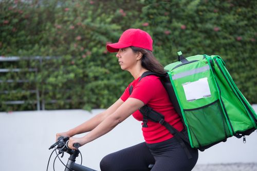 Free A female courier wearing a red cap rides a bicycle with a large green delivery bag outdoors. Stock Photo