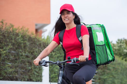 A female courier in a red cap riding a bike with a green insulated bag outdoors.