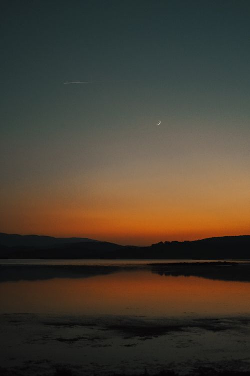 Free Tranquil view of a crescent moon over a calm lake at sunset, reflecting the vibrant orange sky. Stock Photo