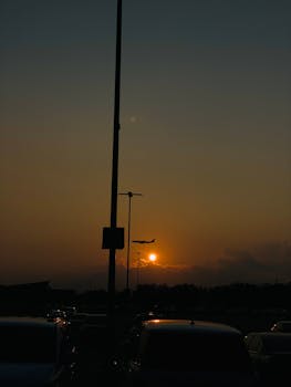 A dramatic sunset scene with a silhouette of a plane landing and cars in a parking lot.