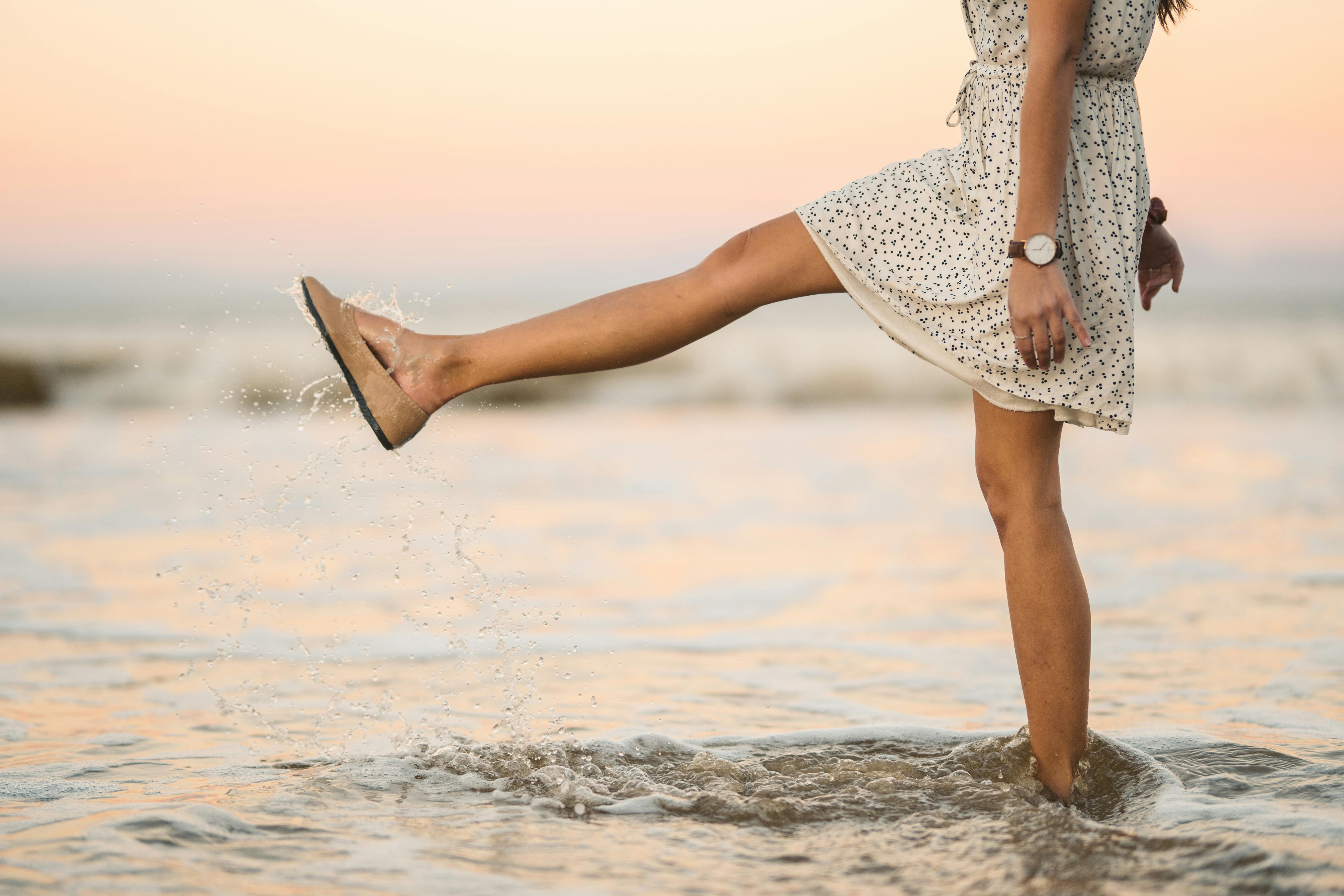 A woman in a sundress kicks water playfully on the beach during a serene sunset.