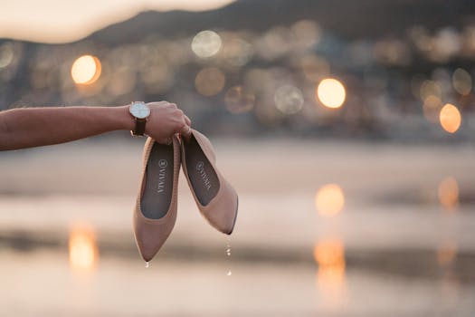 Stylish brown slip-on shoes held by hand at sunset with seaside bokeh background.