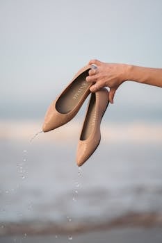 A hand holding stylish brown sandals on a serene beach at sunrise, showcasing simplicity.
