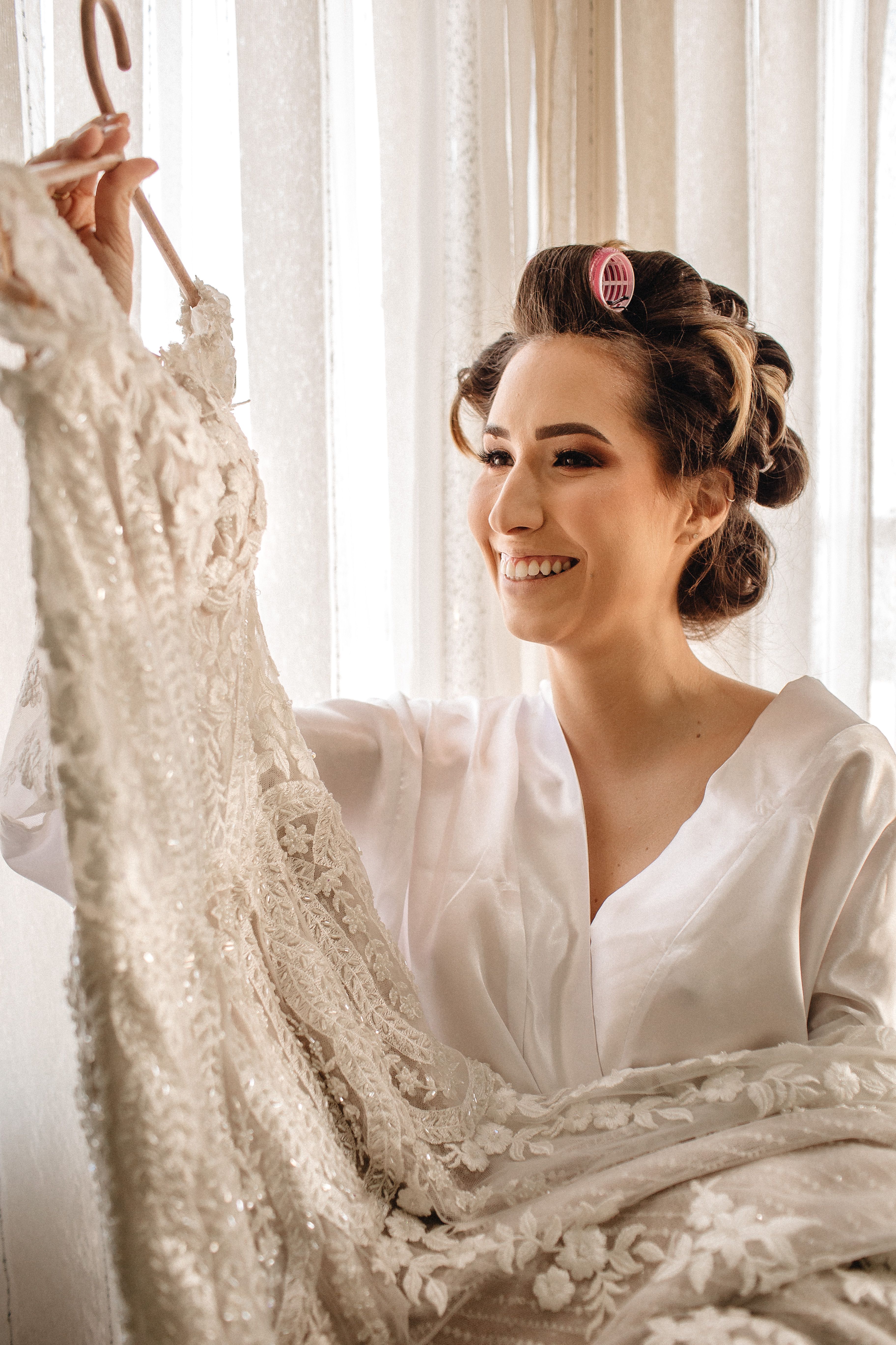 Smiling bride happily admiring her exquisite wedding gown with curlers in her hair.