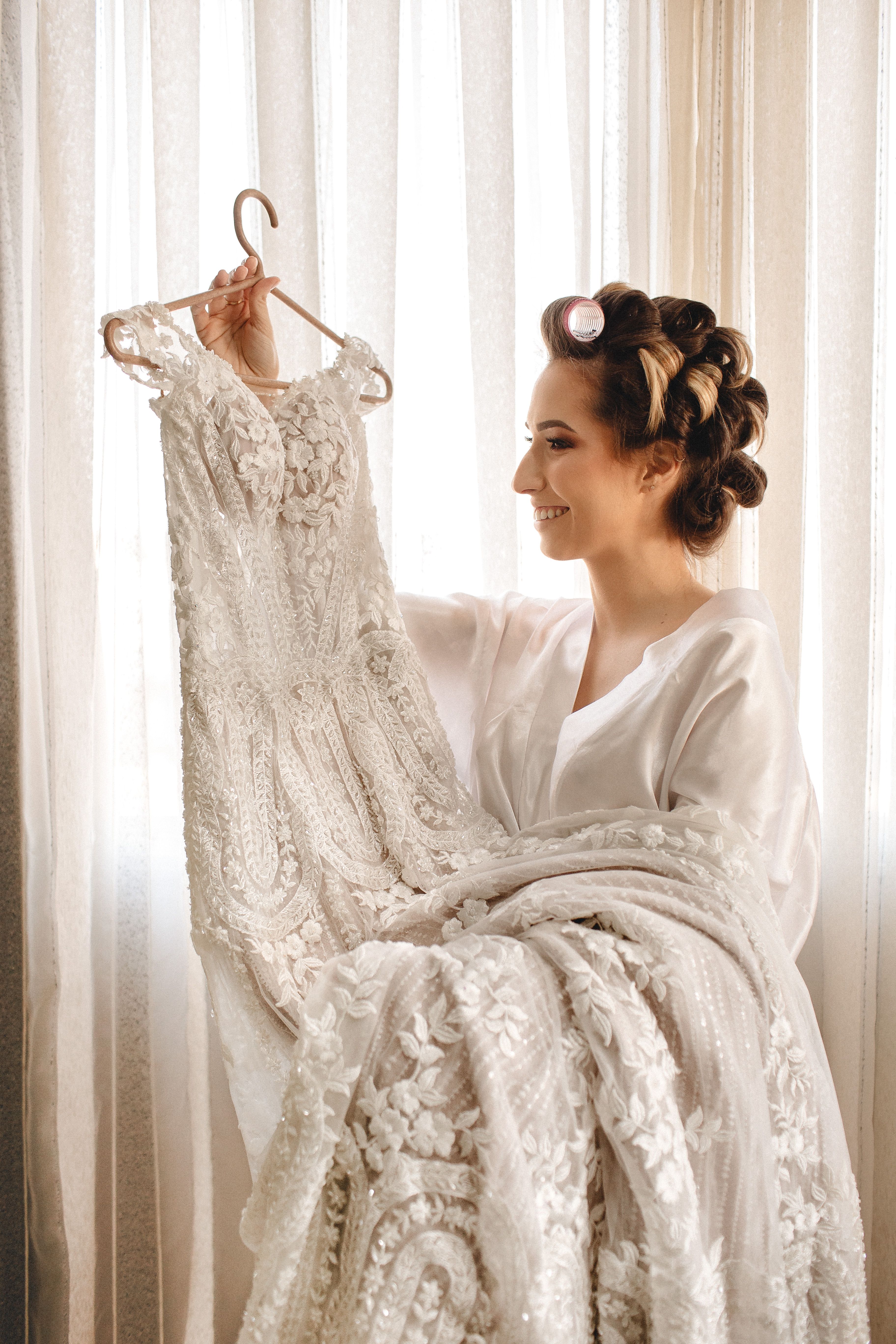 A joyful bride in curlers admires her elegant wedding gown indoors, captured in soft natural light.