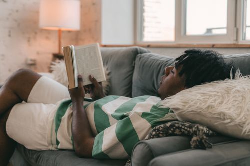 Free A man with dreadlocks lies on a couch reading a book in a cozy indoor setting. Stock Photo