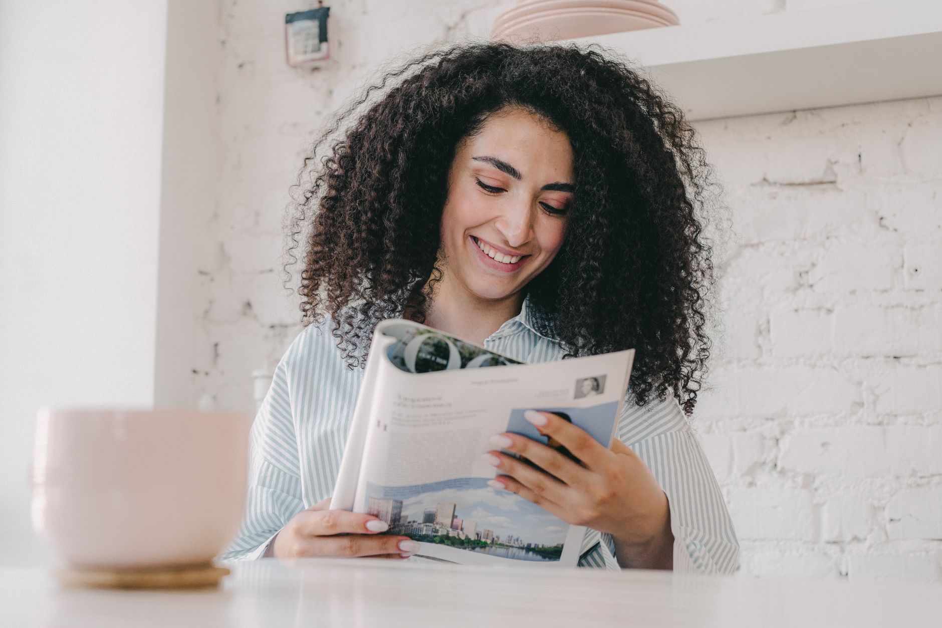 Smiling woman with curly hair enjoying a magazine at home with a ceramic mug on the table.