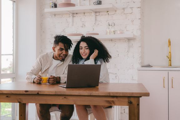 A young couple sits at a wooden kitchen table looking at a laptop together, one holding a glass of orange juice