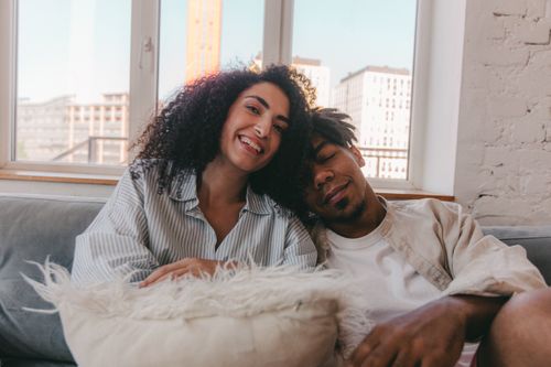 Free A happy couple with curly hair and dreadlocks sitting together on a sofa near a window. Stock Photo