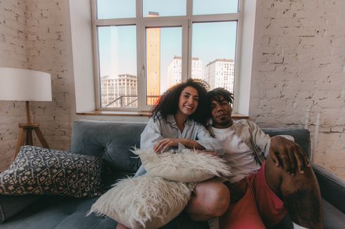 Free Joyful couple sitting together on a sofa in a modern apartment, with city views framing their warm smiles. Stock Photo