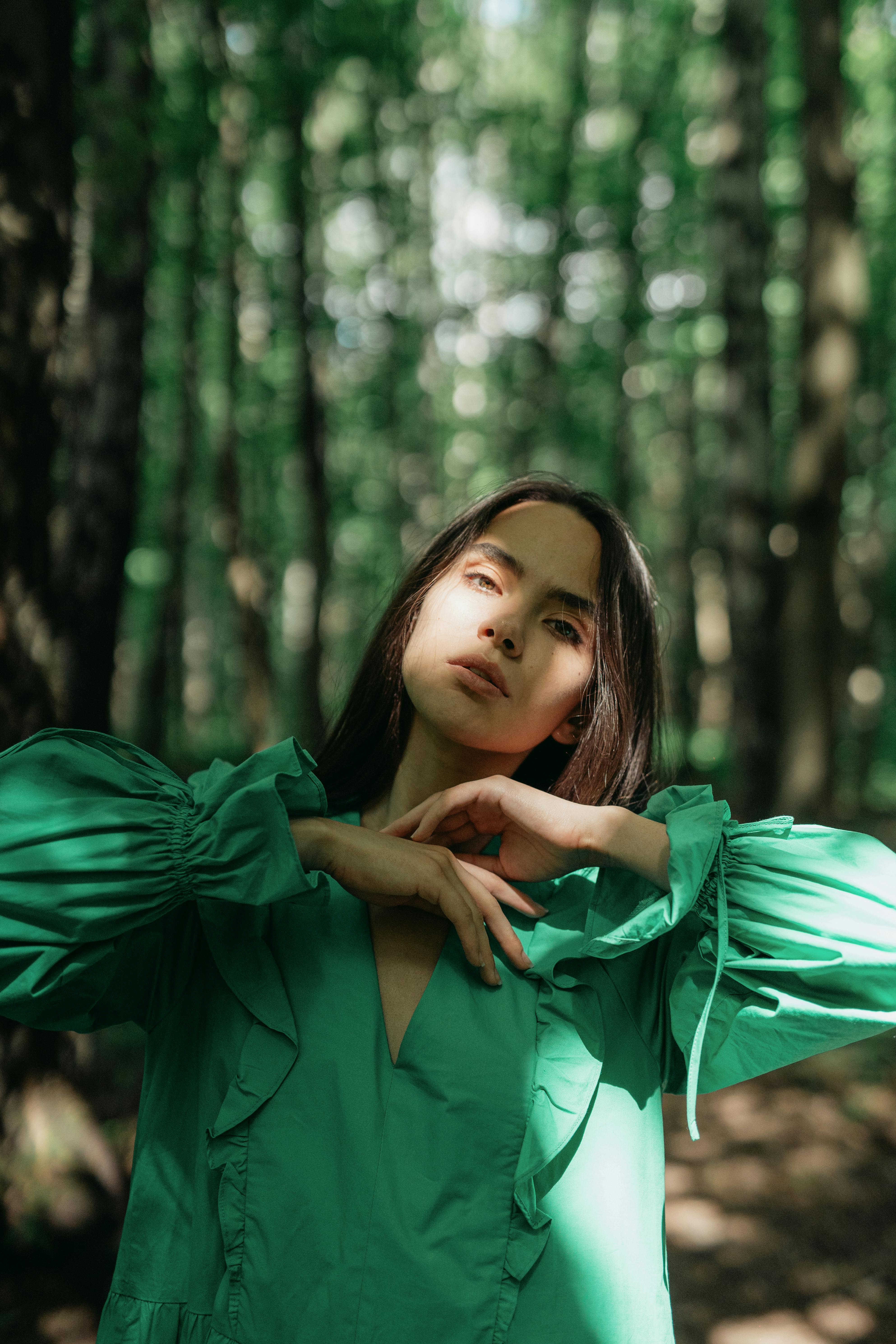 A woman in a green dress poses gracefully in a sunlit forest, creating a serene and natural scene.