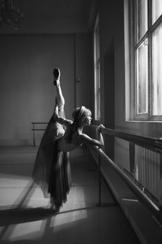 Monochrome photo of a ballerina practicing ballet stretches at the barre indoors.