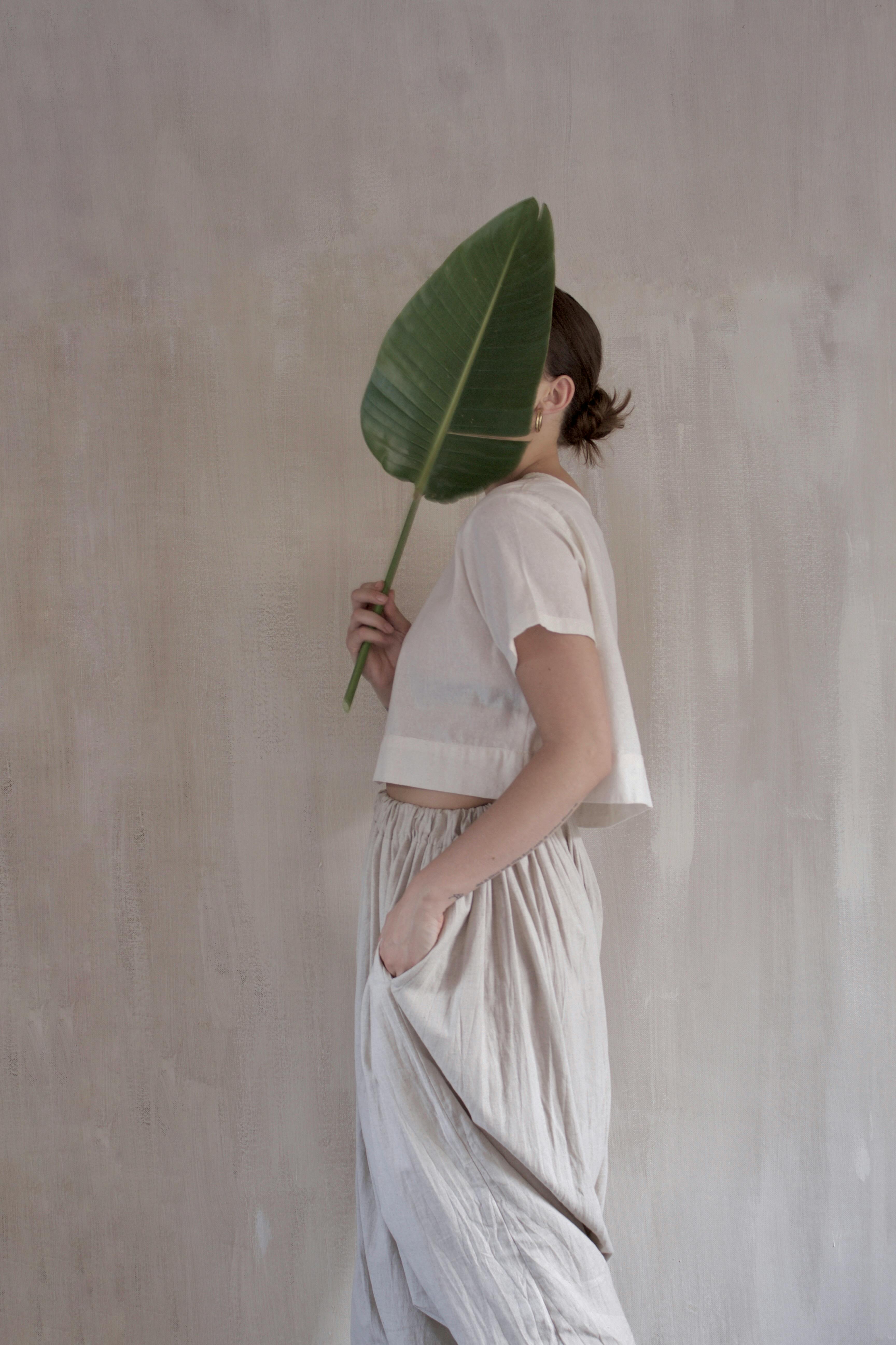Woman holding a large leaf to cover her face, evoking nature and mystery in a minimalist style.