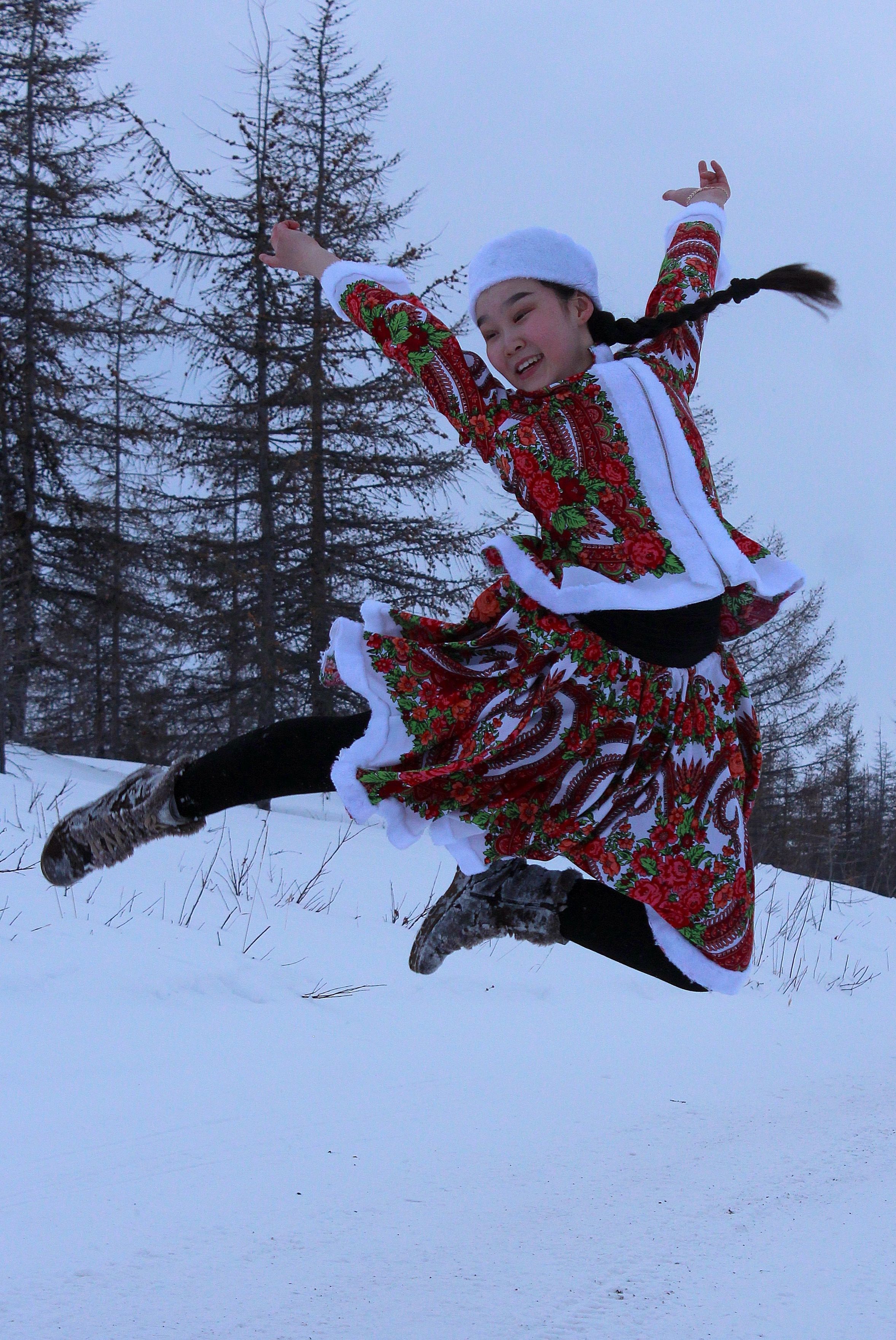 A woman joyfully leaps in snow wearing colorful traditional attire against a winter landscape.