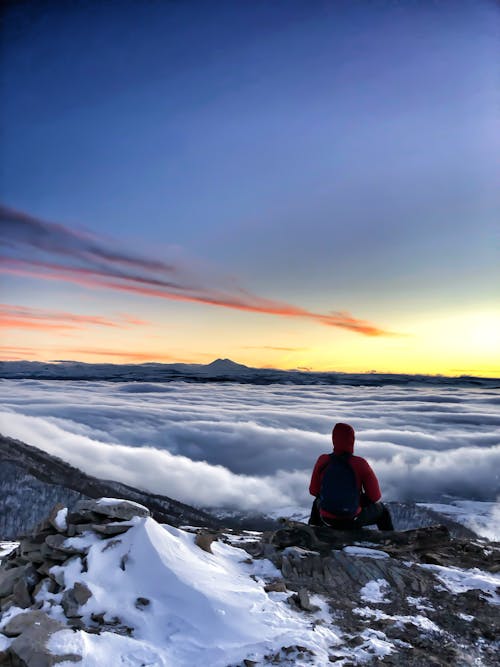 A Person in Red Hoodie Jacket Sitting on a Mountain Peak Covered with Clouds