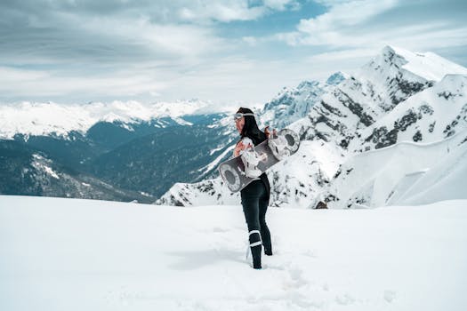 Woman snowboarder in winter mountains, holding gear, surrounded by snowy peaks.