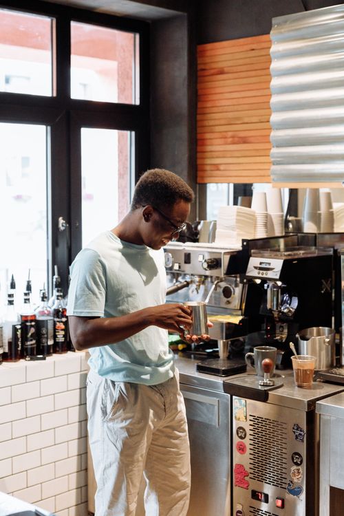 Free Barista in light attire making coffee at espresso machine in a chic, well-lit café. Stock Photo
