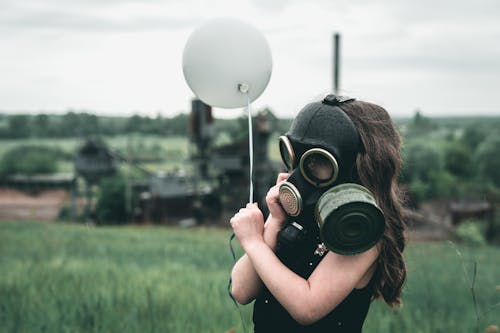 Free A child wearing a gas mask holds a balloon in a field, symbolizing innocence amidst industrial backdrop. Stock Photo