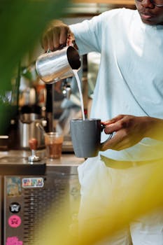 Barista in a white shirt pours milk into a grey mug at a coffee shop, creating latte art.