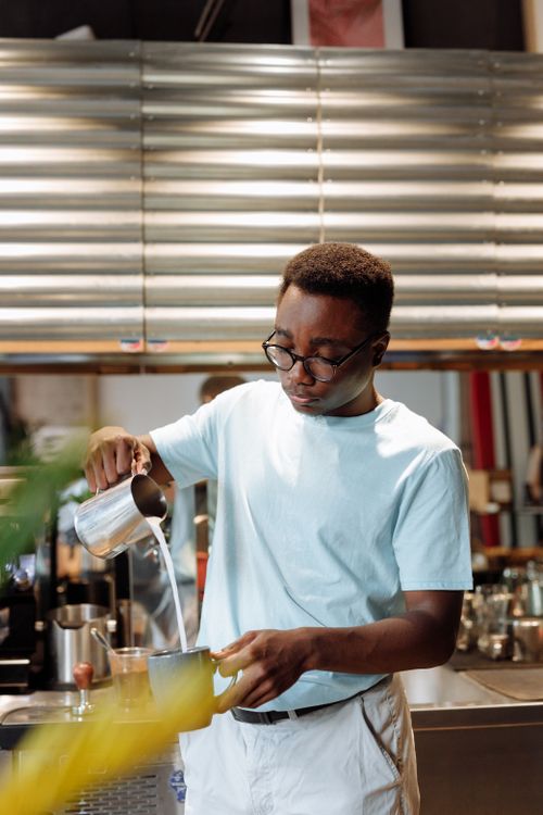 Free Barista pouring milk into coffee at a café counter. Warm, modern indoor setting. Stock Photo