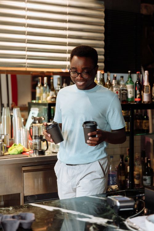 Free Barista in a modern cafe holding two coffee cups, smiling at the counter. Stock Photo