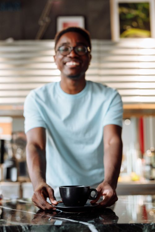 Free Friendly barista presenting a cup of coffee at a modern café counter, creating a welcoming atmosphere. Stock Photo