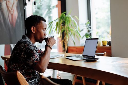 Free A man enjoys coffee while working on a laptop at a wooden table with a plant nearby. Stock Photo