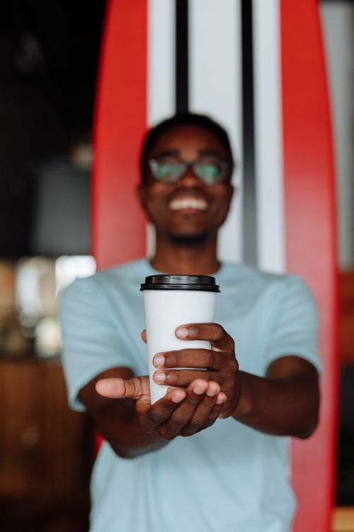 Free Cheerful man with eyeglasses offers a coffee cup indoors, creating a warm and inviting atmosphere. Stock Photo