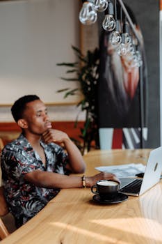 A thoughtful man working at a café table with a laptop and coffee, under stylish lighting.