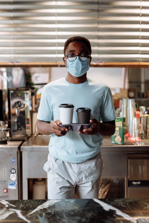 Free Man with face mask and glasses holding coffee cups in a modern cafe indoors. Stock Photo