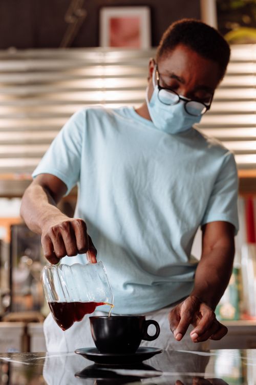 Free Barista wearing mask pouring coffee into cup in a cafe, emphasizing pandemic safety. Stock Photo