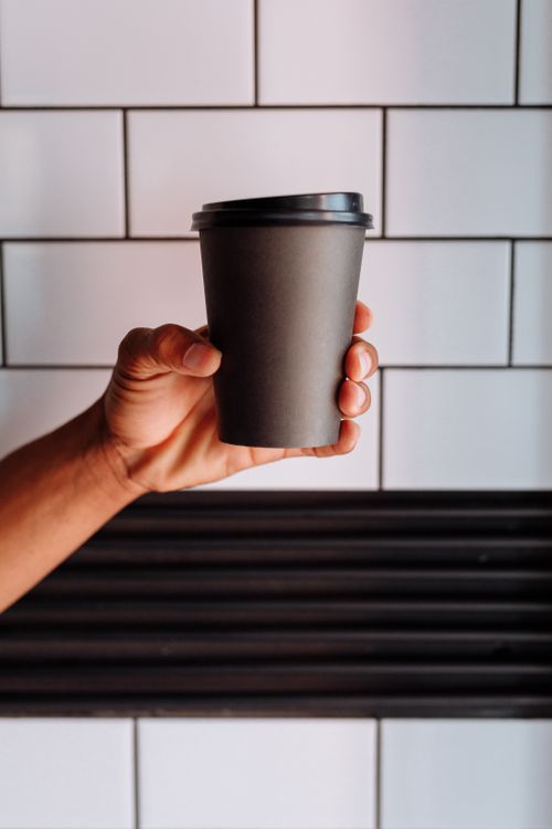 Free A hand holds a black disposable coffee cup against a modern tiled background, offering a stylish and minimalist vibe. Stock Photo