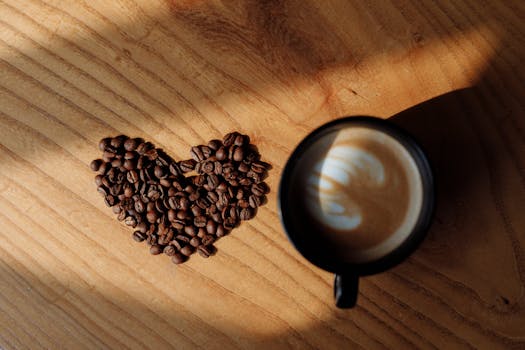 A heart-shaped arrangement of coffee beans next to a latte on a wooden table.