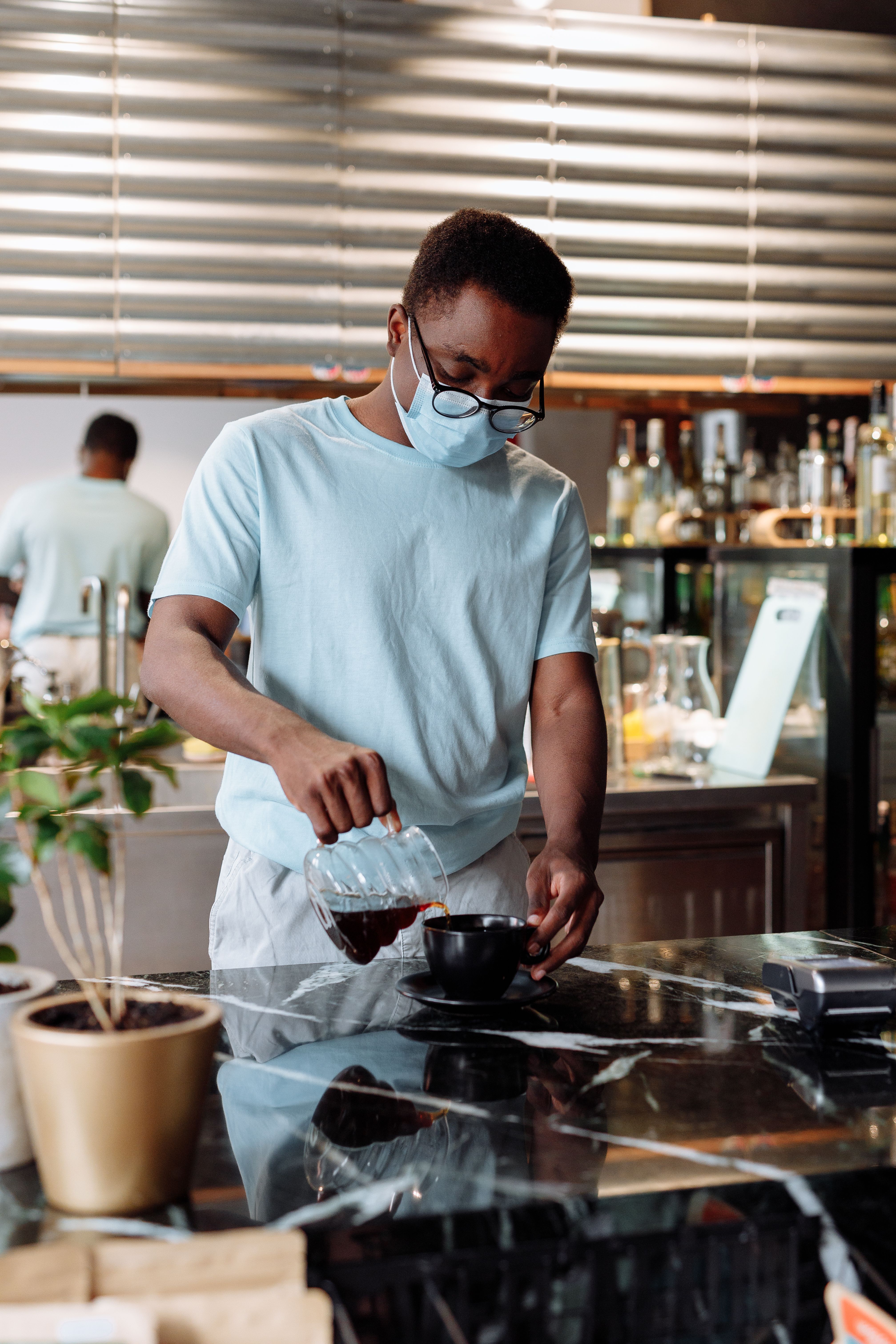 A barista wearing a mask prepares pour-over coffee in a modern cafe setting.