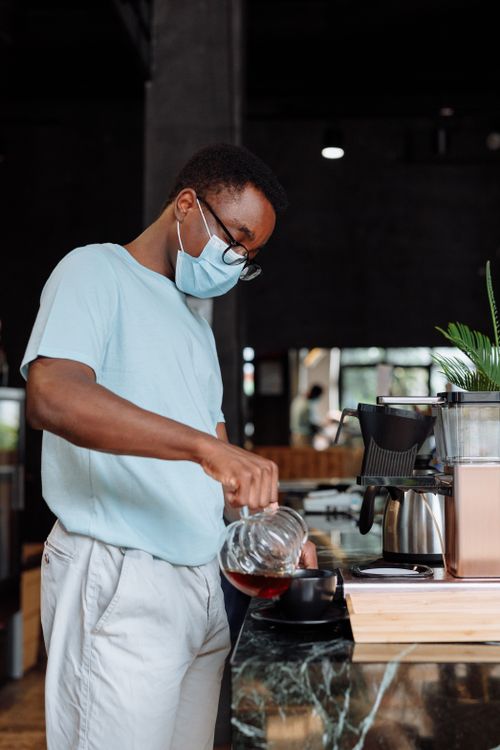 Free A masked barista pours coffee while working inside a modern café, creating a fresh brew. Stock Photo