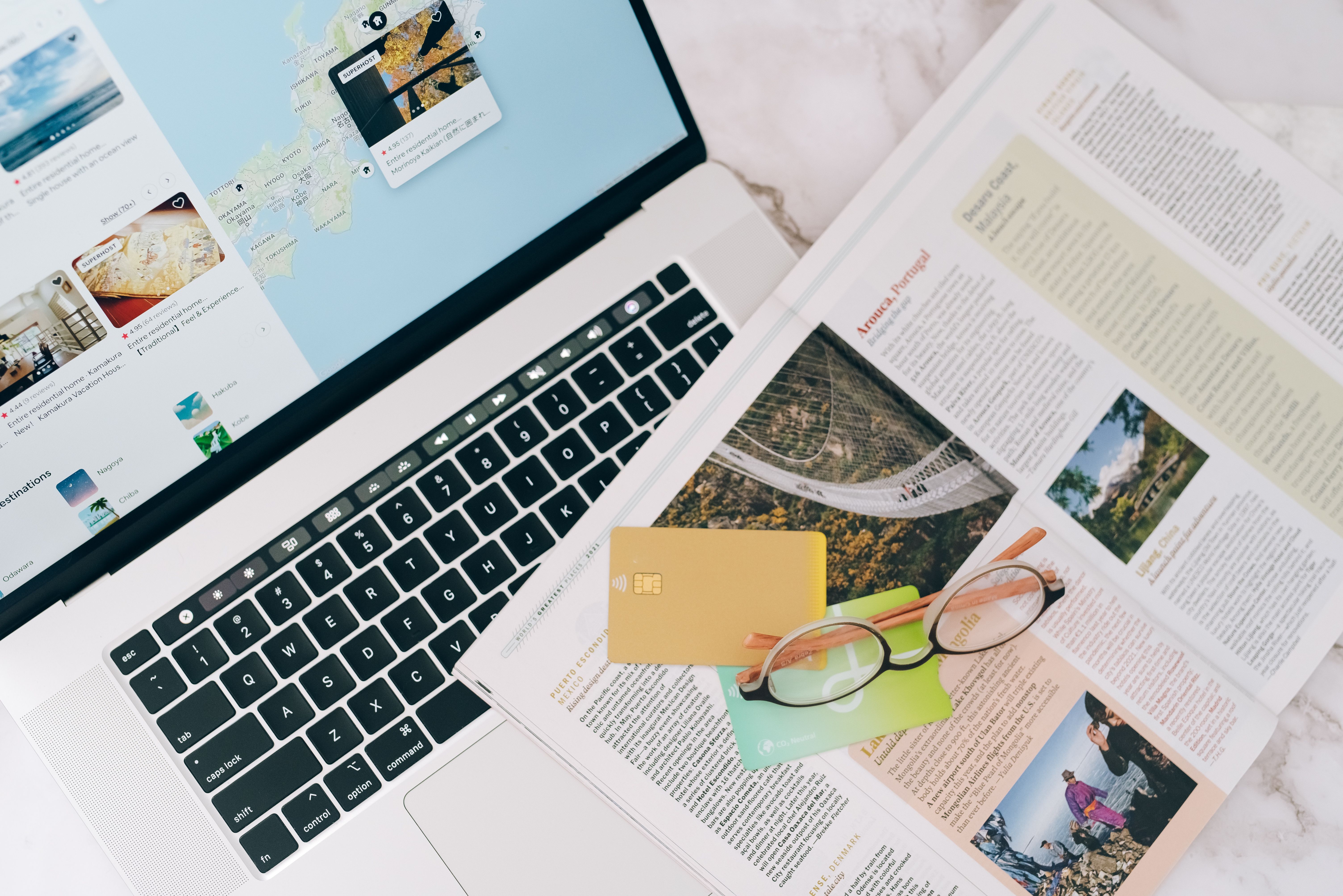 Laptop displaying a travel map beside an open magazine, bank card and glasses on a table