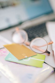 Blurred close-up of credit cards, eyeglasses, and laptop on a desk, evoking online shopping theme.