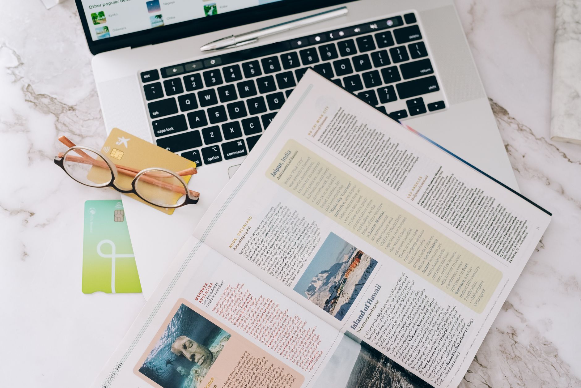 Flat lay image featuring a laptop, open magazine, credit cards, and eyeglasses on a marble table.