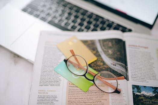 Overhead view of reading glasses, credit cards, and a magazine with a blurred laptop in the background.