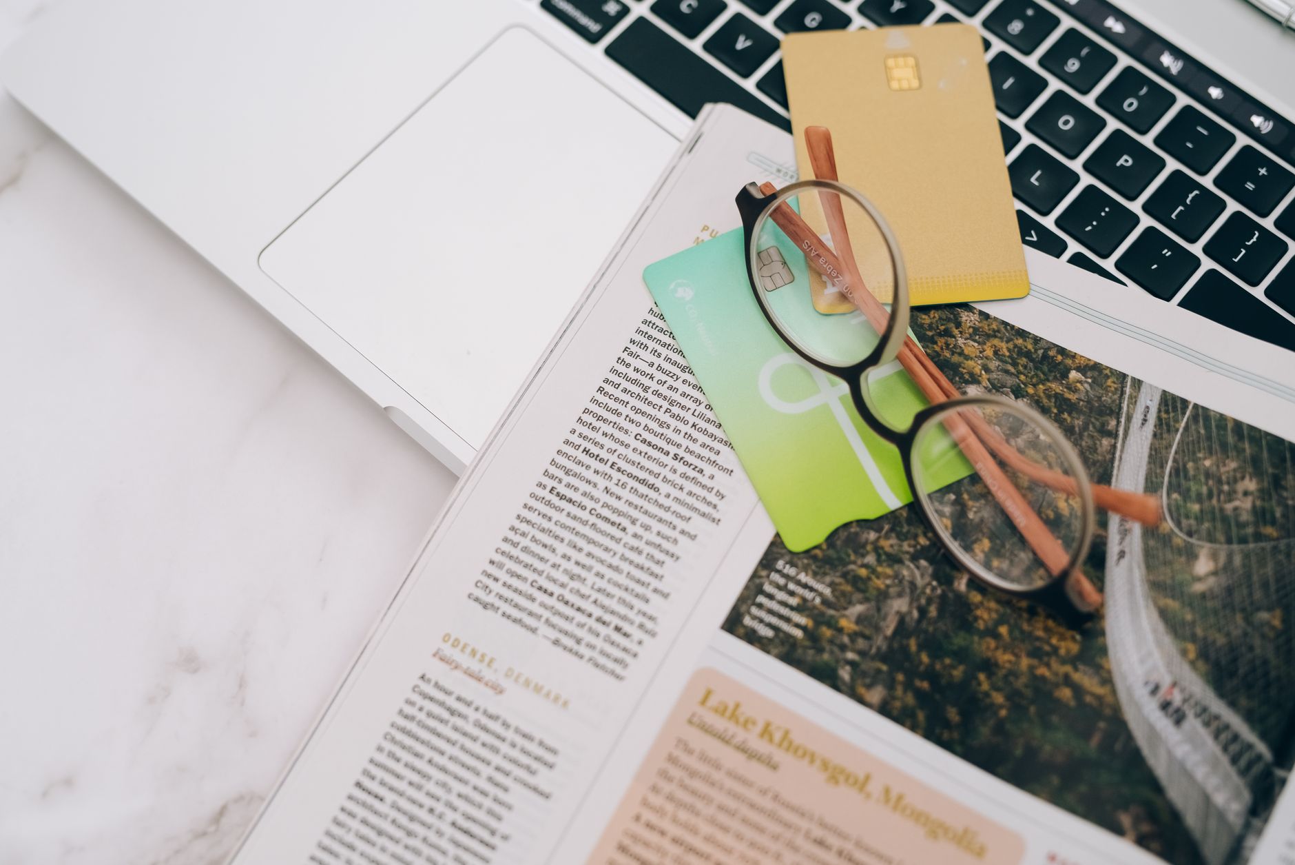 Eyeglasses resting on a magazine beside a laptop and credit cards, indoors.