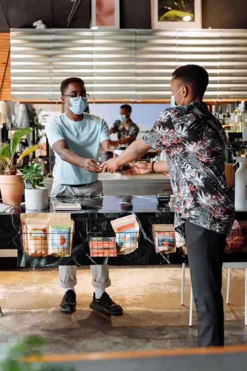 Free Two men wearing masks interact in a stylish cafe setting, exchanging items over a counter. Stock Photo