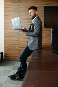 A man in a suit jacket stands in an office, holding a laptop and working.