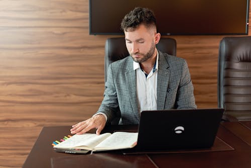 Man in Gray Suit Jacket Sitting Behind His Desk Looking On A Notebook