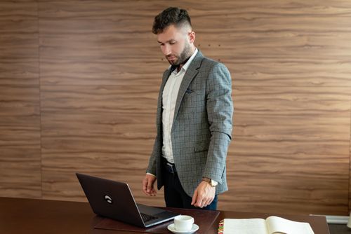 Free Businessman in gray blazer stands working on laptop at office desk. Stock Photo