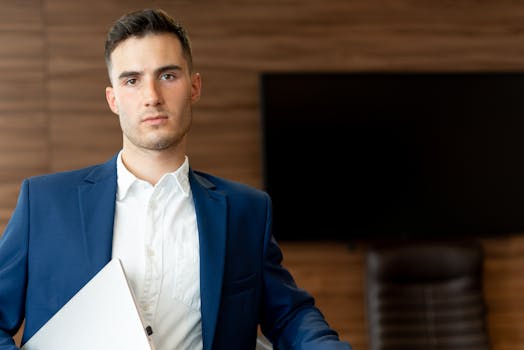 Confident young businessman in a blue suit, holding a laptop in an office setting.