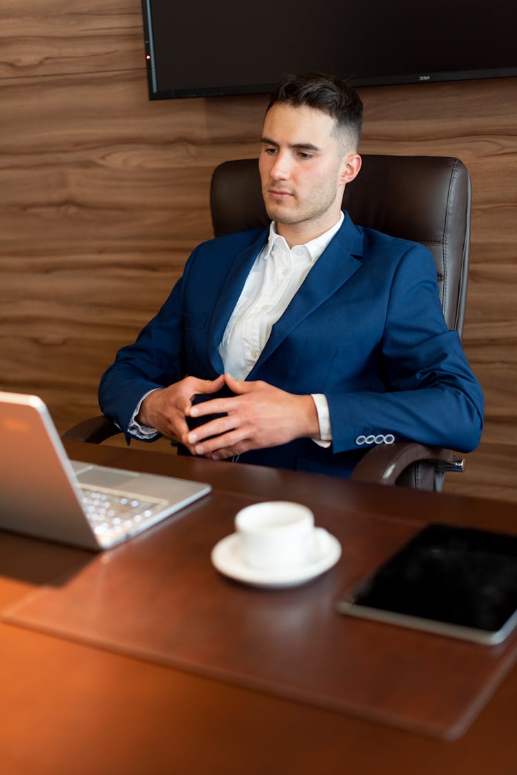 A Man In Blue Suit Sitting On The Chair While Looking At The Laptop