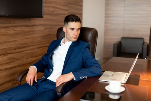 Free Confident businessman in blue suit seated at a desk with laptop, contemplating in office setting. Stock Photo