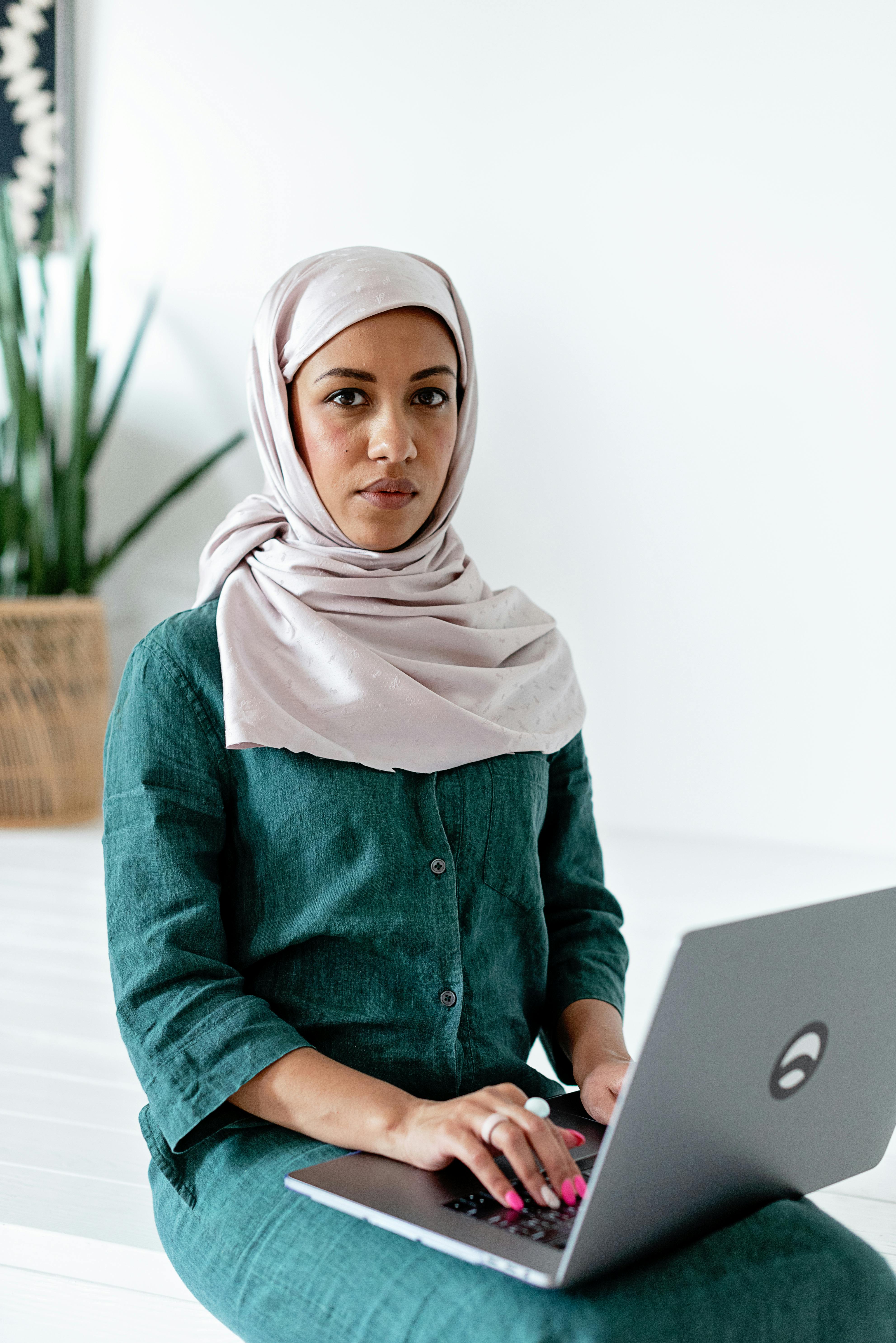 Serious woman in a hijab sitting indoors, working on a laptop. Professional and focused atmosphere.