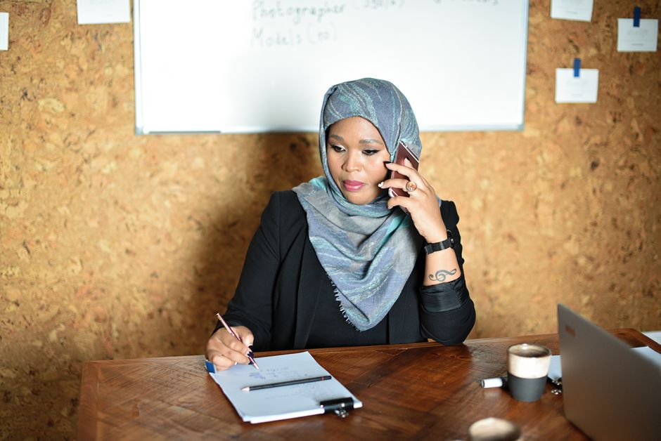 Businesswoman in a hijab working and talking on phone, taking notes in office with a whiteboard behind.