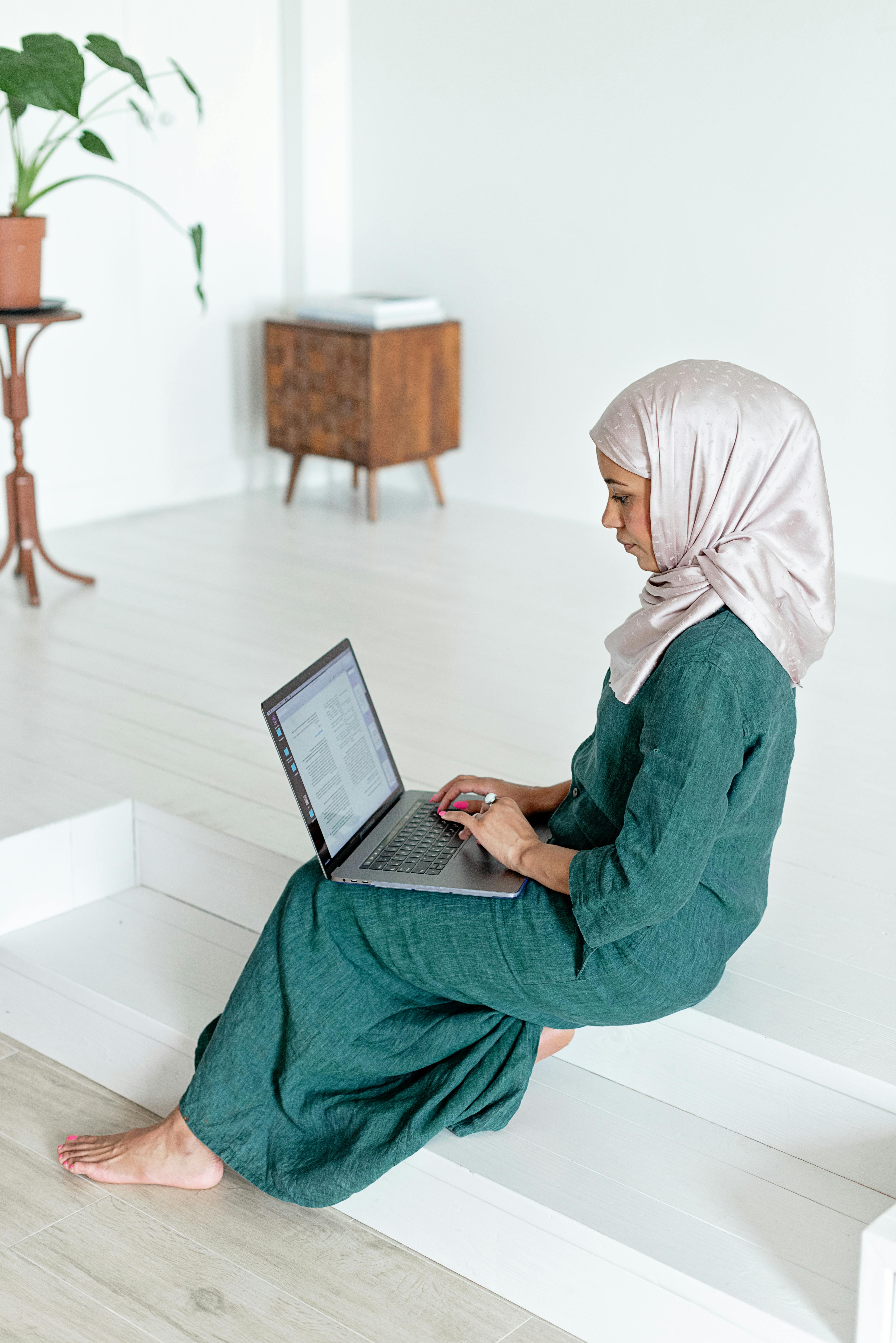 Woman wearing hijab and green dress works on laptop while sitting on indoor wooden steps.